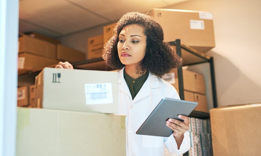 A woman wearing a white doctor's jacket looks closely at a stack of cardboard boxes while holding a tablet.