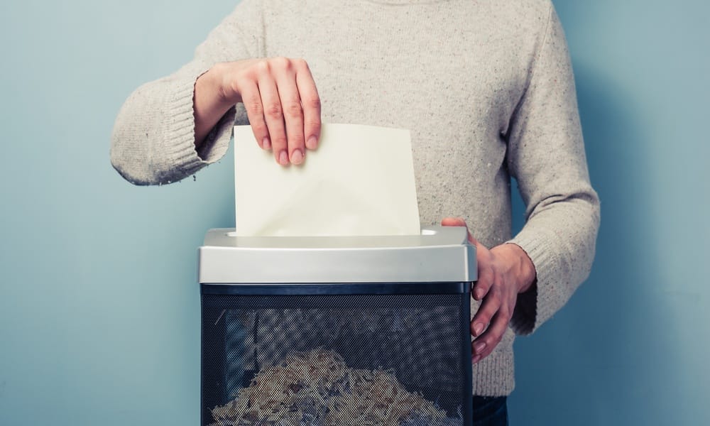 A person wearing a gray sweater is pushing a white piece of paper into a black and gray shredder.