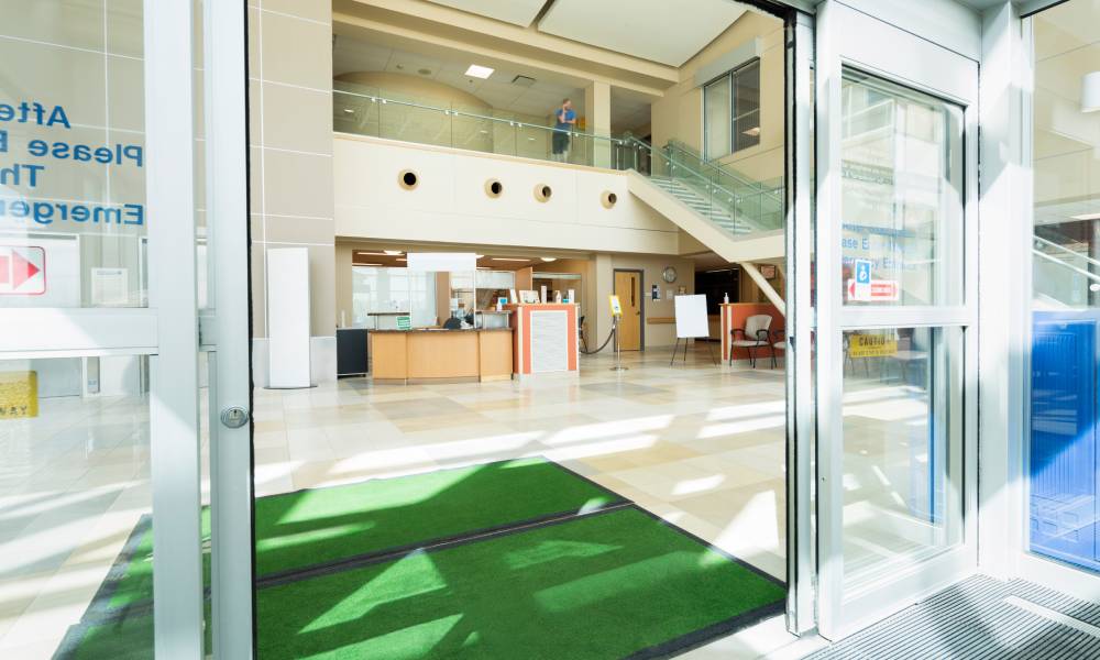 Green doormats on clean floors at the entrance of a facility. The facility has open, automated doors.