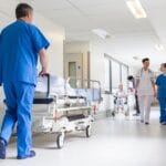 A hospital hallway with doctors and nurses walking through. There's a doctor pushing an empty bed on the left.