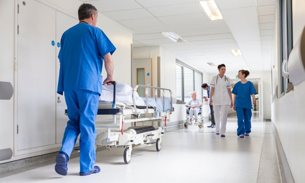 A hospital hallway with doctors and nurses walking through. There's a doctor pushing an empty bed on the left.