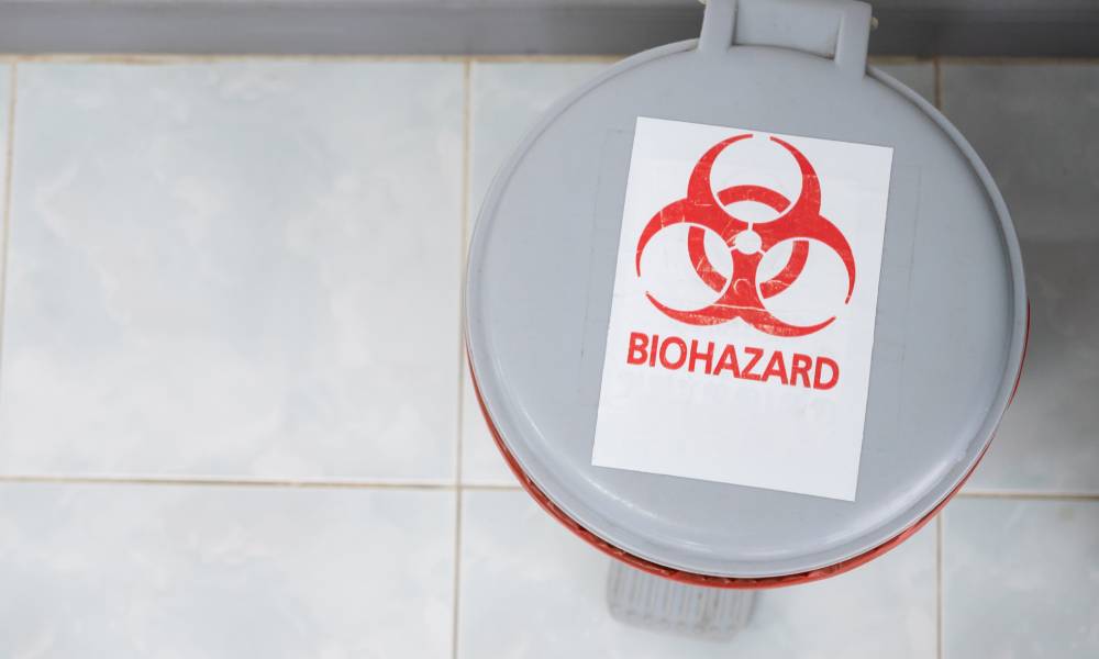 The top of a small, gray trash bin on the floor with "biohazard" written in red on the lid with a matching red symbol.