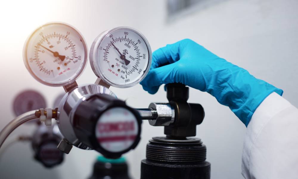 A person wearing gloves and turning the top valve on a gas cylinder with two large round pressure gauges.