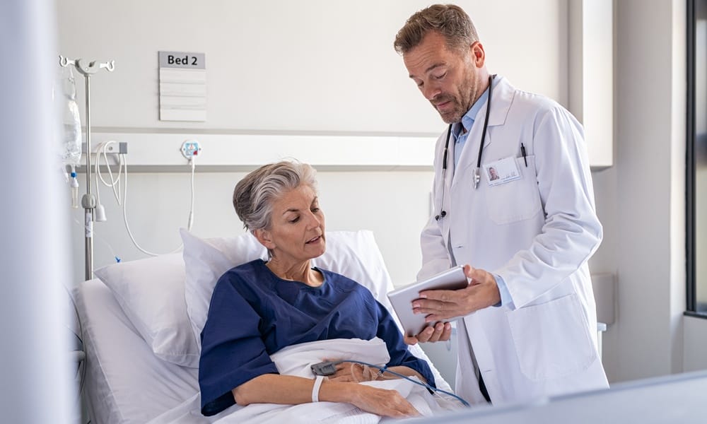 A medical professional in a white coat speaking with a mature patient and showing them a silver tablet.