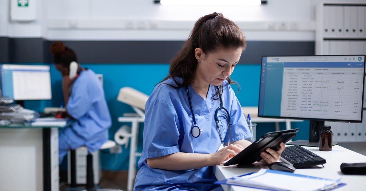 A woman in scrubs uses an electronic tablet while sitting at a desk in front of a computer in a healthcare setting.