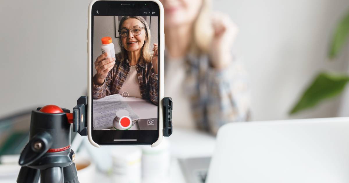 A mature person sitting across from a recording smartphone on a tripod and holding up a white pill bottle.