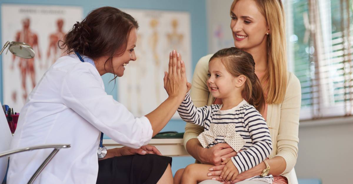 A person wearing a doctor's lab coat and stethoscope high-fives a younger kid sitting on another woman's lap.