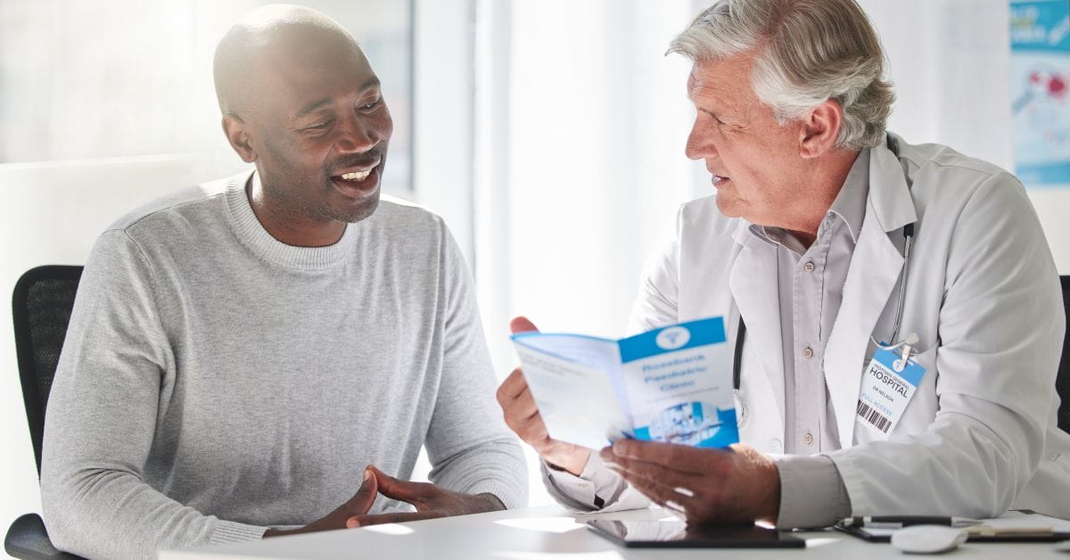 A doctor and his patient sitting next to each other. The doctor holds a medical pamphlet while talking to the patient.