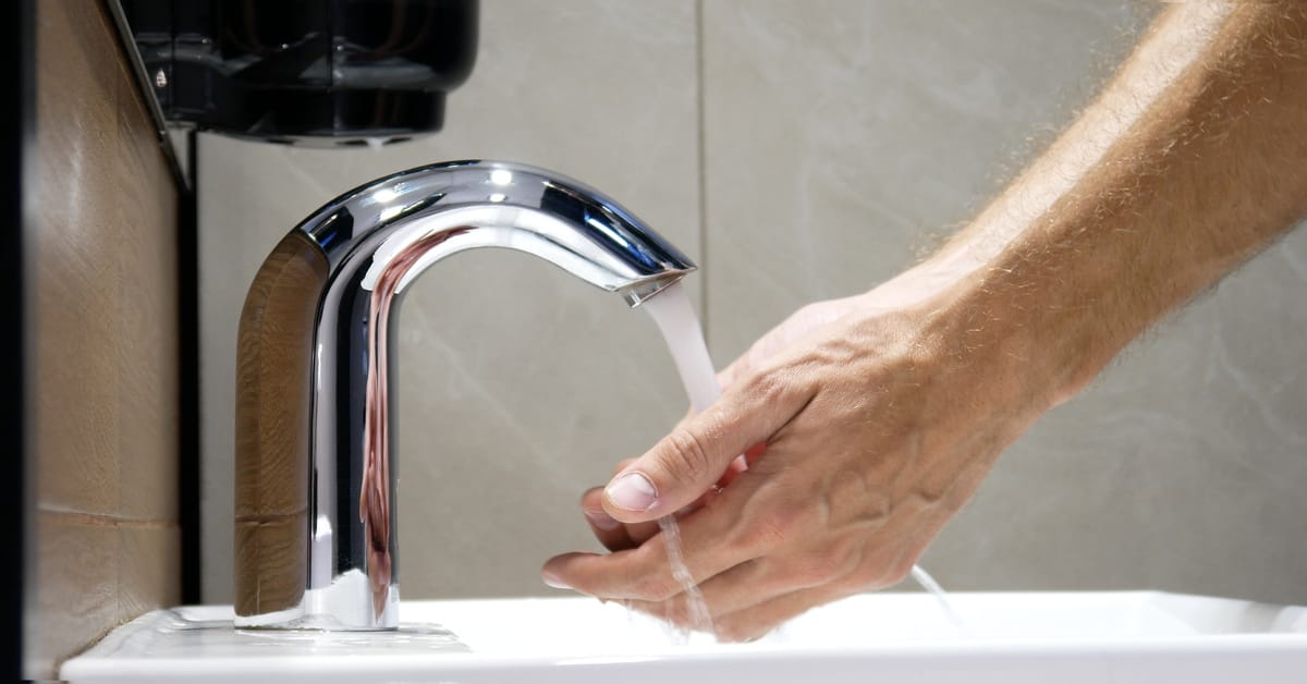 A man is washing his hands at a bathroom sink. Water pours from the touchless faucet while a soap dispenser is on the wall.
