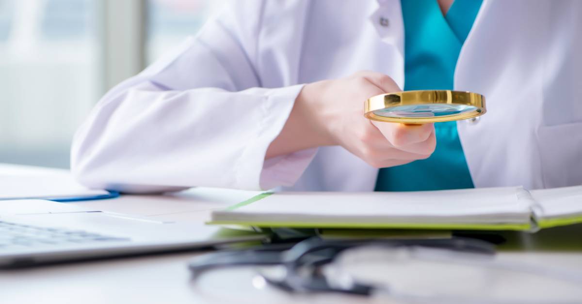 A person in teal scrubs and a white lab coat holds a gold magnifying glass over an open spiral-bound notebook.