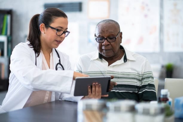An elderly African American man wearing glasses sits next to a female doctor.  The doctor is wearing glasses, a white lab coat and has a stethoscope around her neck.  She is discussing the man's medical chart