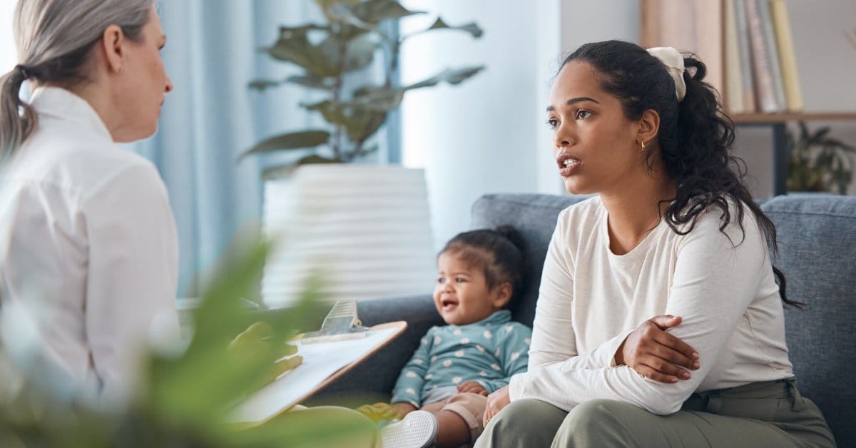A woman in professional attire, holding a clipboard, is sitting across from a woman and her child. The patient is concerned.