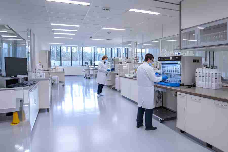 Two scientists wearing white lab coats and facemasks, standing in front of a large workspace table with equipment.