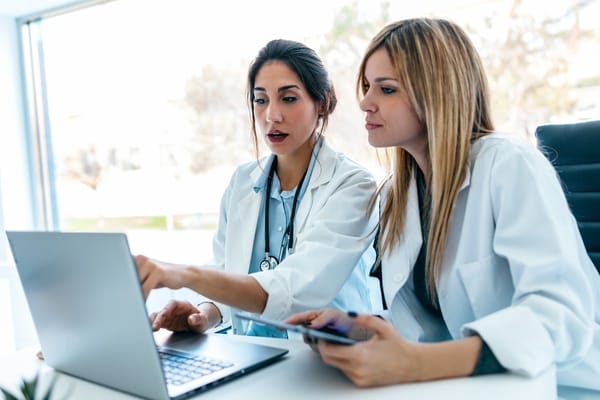 Two women wearing white jackets and stethoscopes are sitting in a brightly lit office and looking at a laptop.