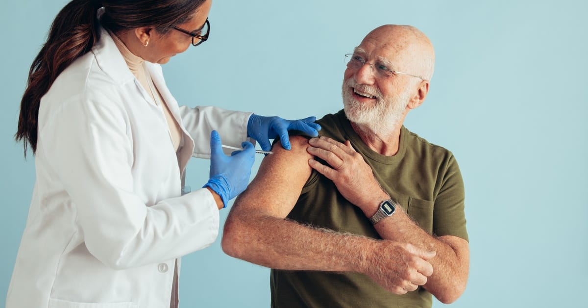 A doctor wears a white lab coat and administers a vaccine in the arm of a smiling senior man against a blue background.