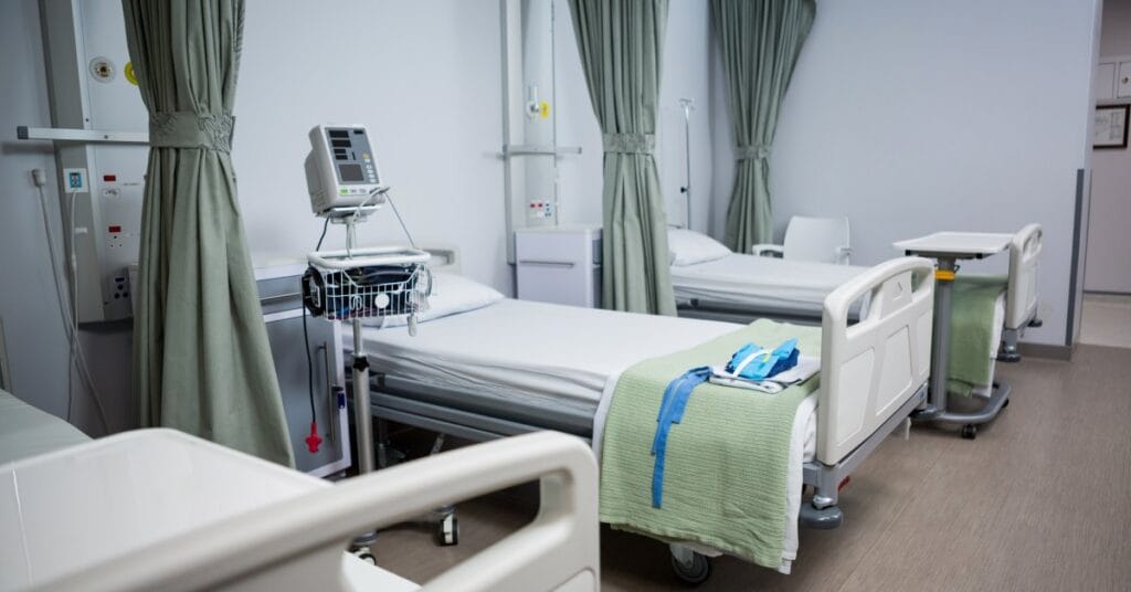 A hospital room with three patient beds set up and neatly made. The green curtains are pulled back with a tie.