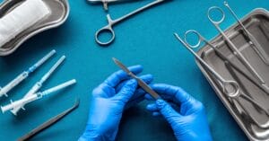 A doctor wearing blue gloves holds a scalpel on a blue table. The table also contains other surgical equipment.
