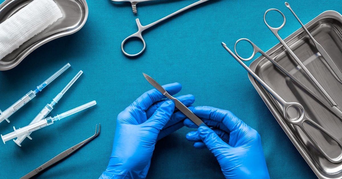 A doctor wearing blue gloves holds a scalpel on a blue table. The table also contains other surgical equipment.
