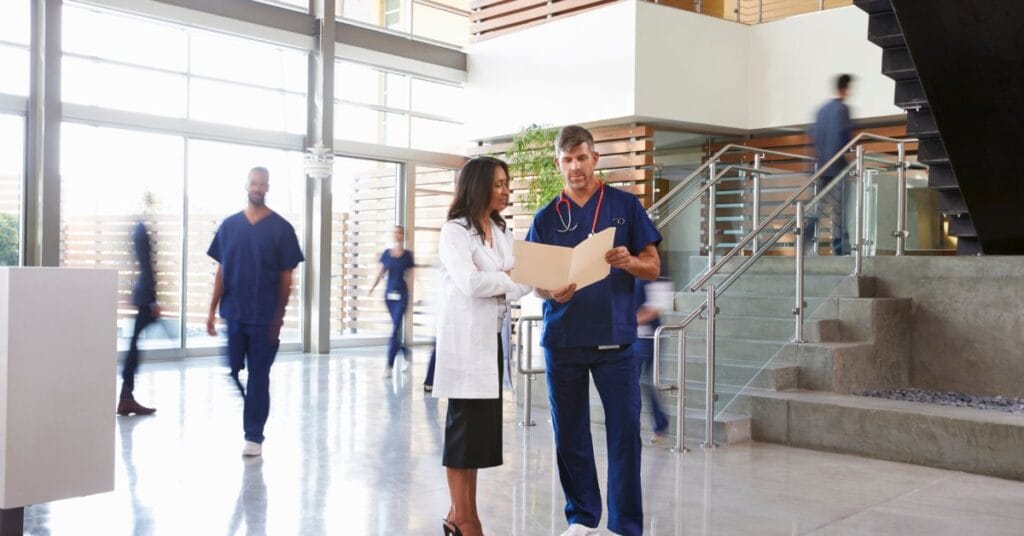 Two healthcare workers, one in scrubs and one in a white coat, talk in a facility lobby while reviewing paperwork.
