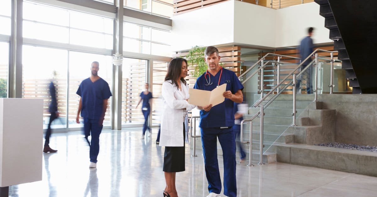 Two healthcare workers, one in scrubs and one in a white coat, talk in a facility lobby while reviewing paperwork.