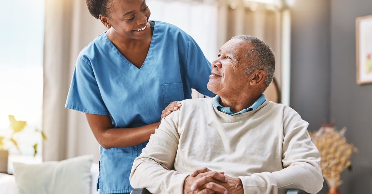A nurse stands behind an older gentleman in a wheelchair. She has her hand on his shoulder, and they smile at each other.