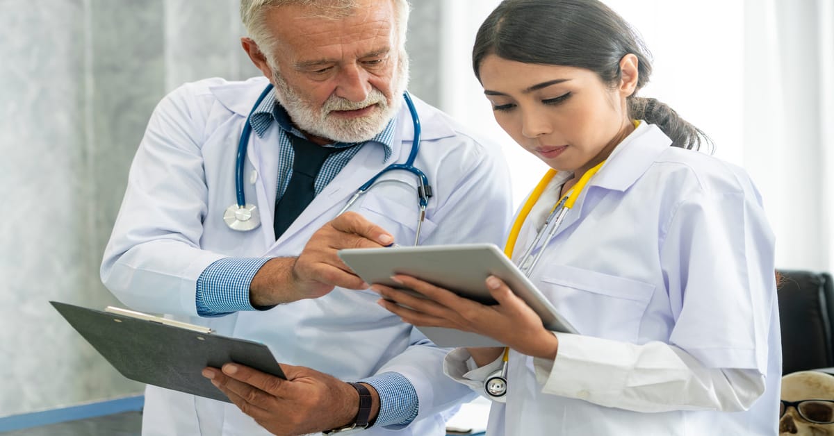 An older doctor is holds a clipboard and points toward a tablet held by a female doctor standing right beside him.