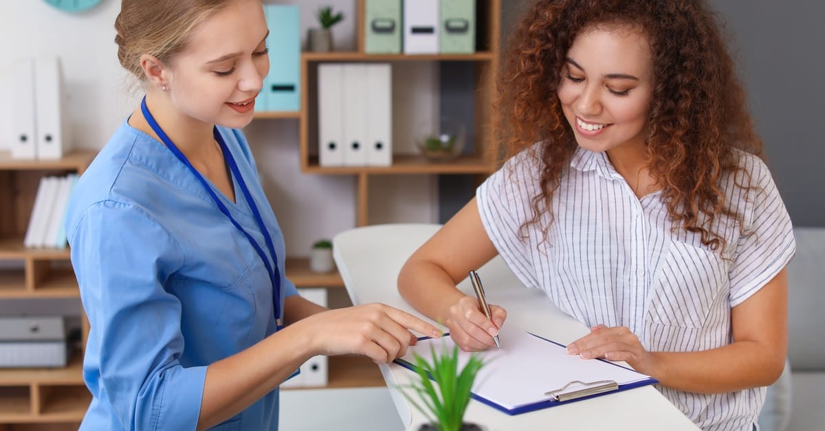 A young female receptionist wearing blue scrubs standing behind a desk with another woman signing a document.