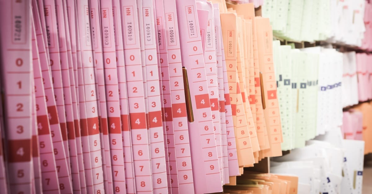 Pink, orange, and white folders are stacked upright on three shelves in a medical office. The files fill the frame.