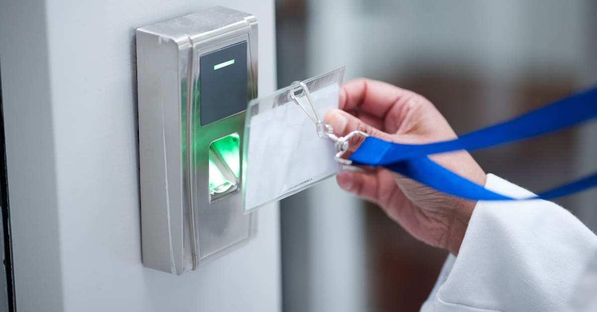 A medical professional in a white lab coat lifting a badge on a blue lanyard toward a reader to gain access to an area.