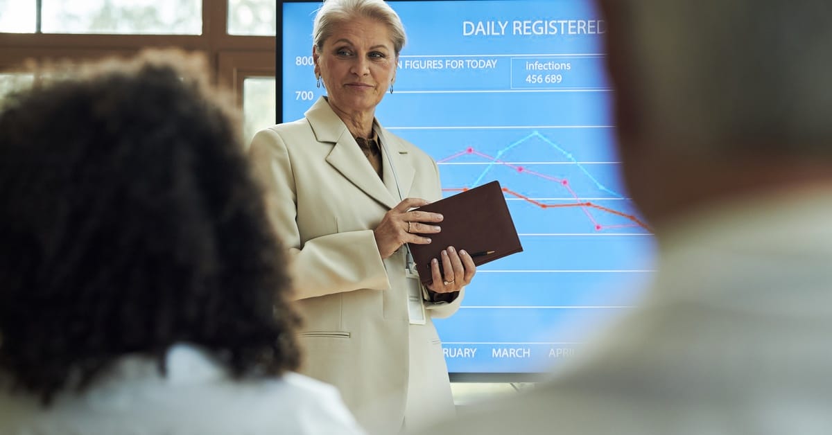 A female executive in a cream suit stands in front of a presentation screen, looking to her audience.