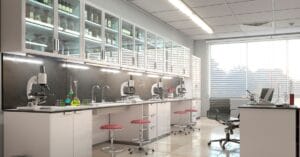 A bright laboratory with microscopes on a long counter, glass cabinets of labware, test tubes, and stools.